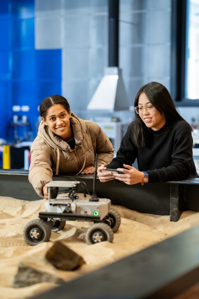 2 students in a robotics lab, controlling a remote control buggie on a sand platform