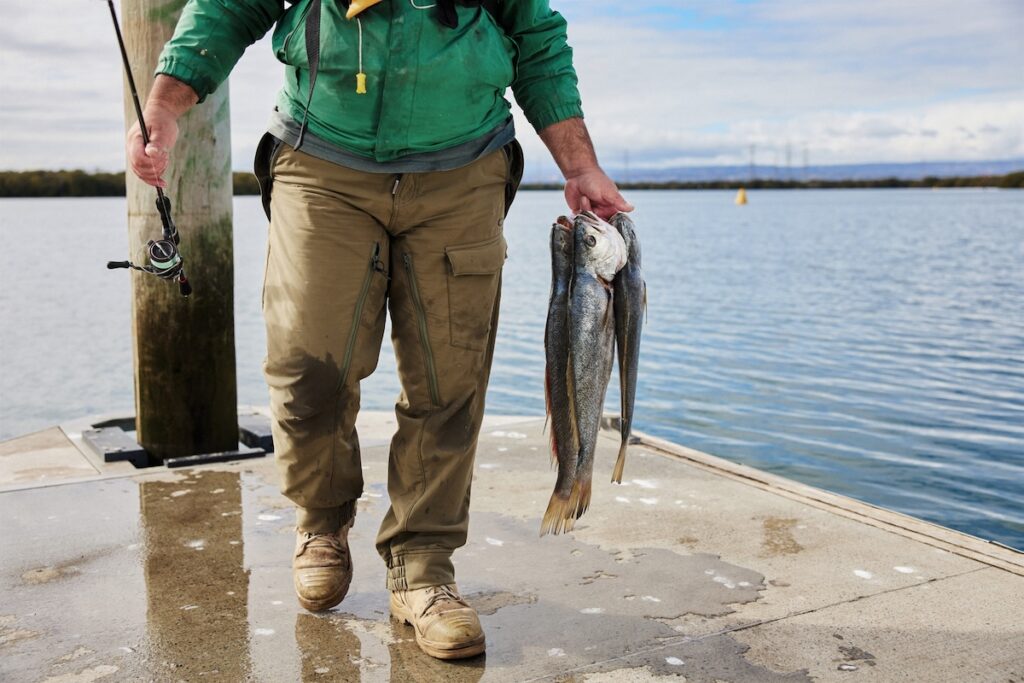 A man walking on a jetty holding a fishing rod in one hand and a few fish in the other
