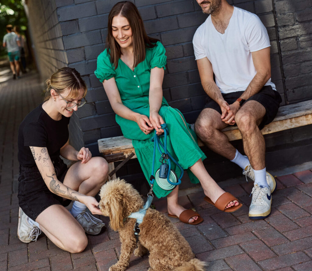 A waitress kneeling down to pat a dog, owned by two people sitting outside on a bench