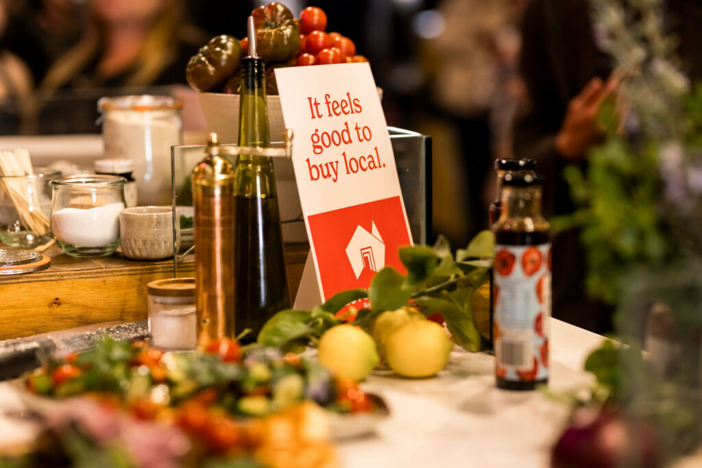 A close up image of a table setting inclusive of decorative leaves, fruits and condiments with a sign that says 'it feels good to buy local'