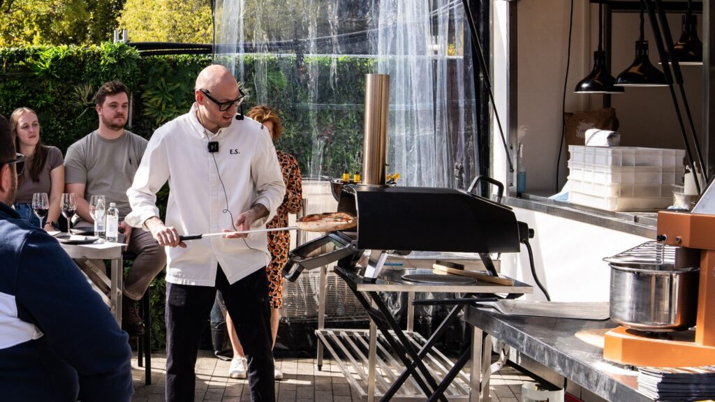Enrico placing pizza into an outdoor oven whilst attendees at surrounding tables watch
