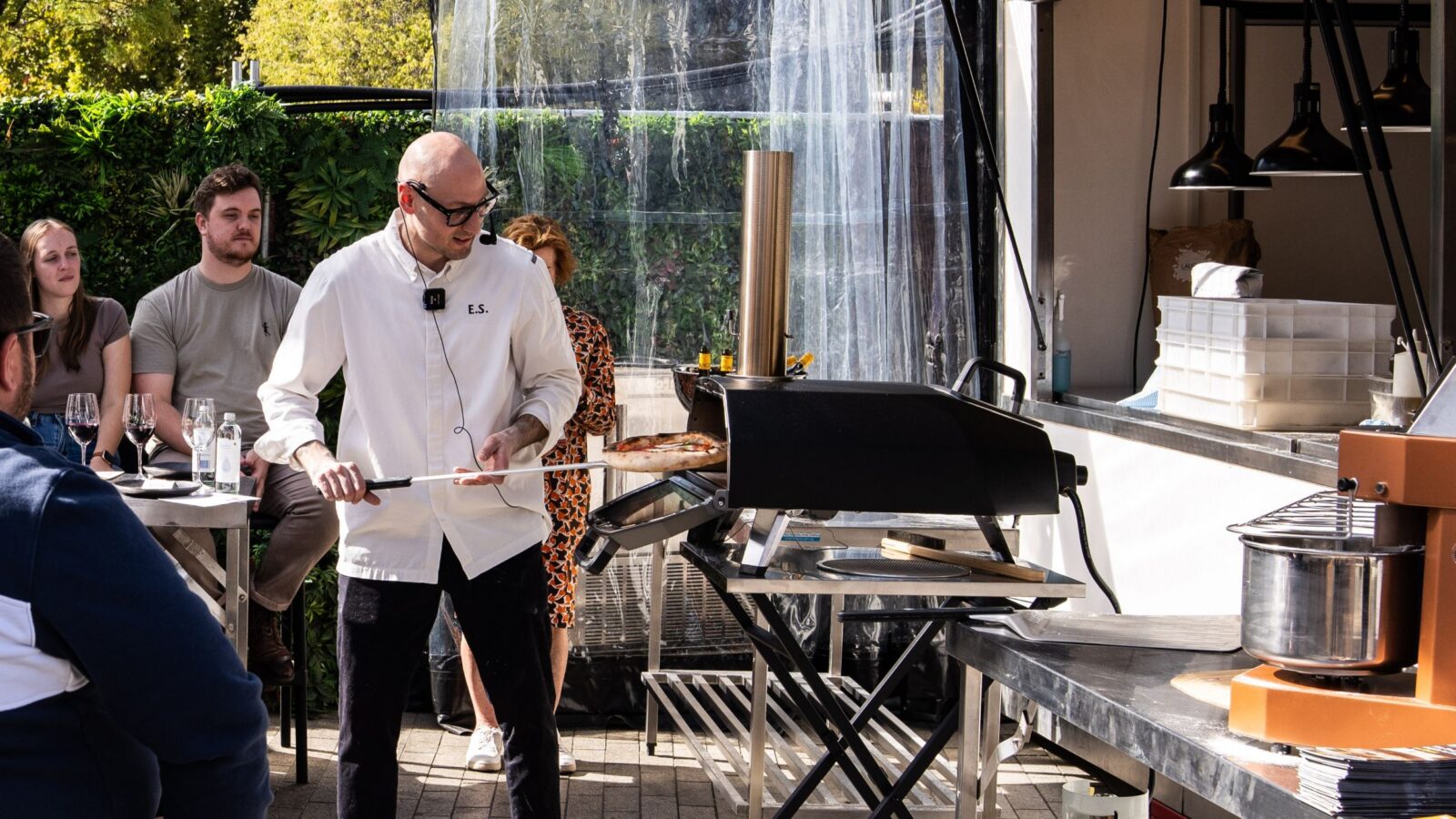 Enrico placing pizza into an outdoor oven whilst attendees at surrounding tables watch