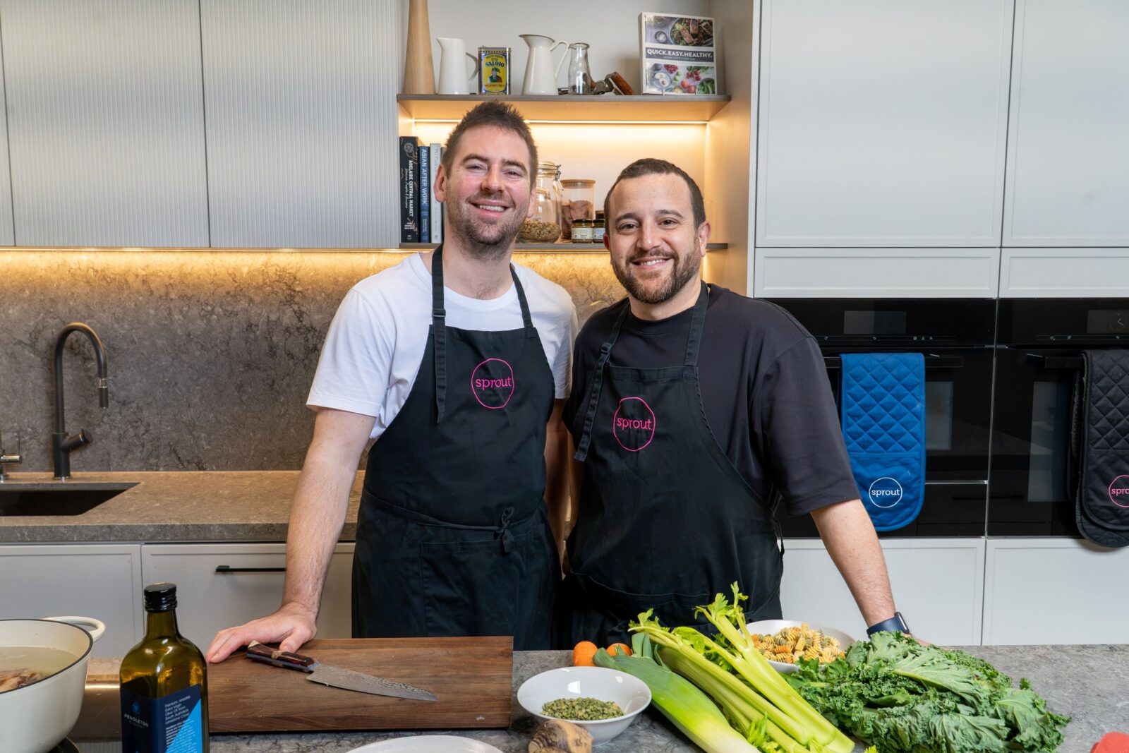 Callum and Themis standing behind a kitchen counter full of fresh vegetables and a cutting board whilst smiling to camera