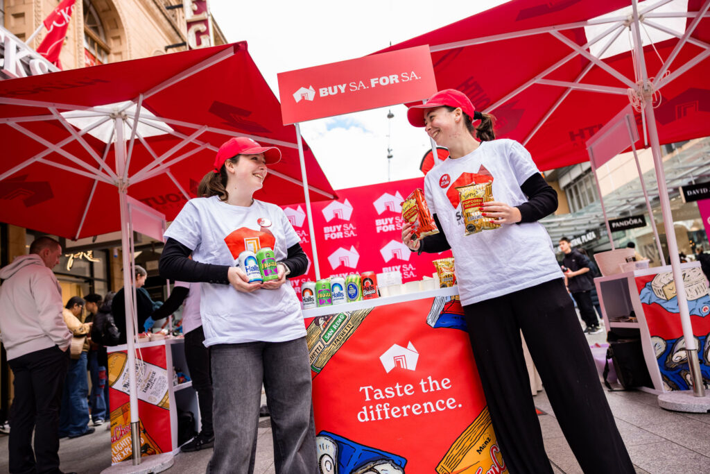 They Buy SA stall in Rundle mall with two people holding up cans of softdrink and packets of popcorn whilst smiling towards each other